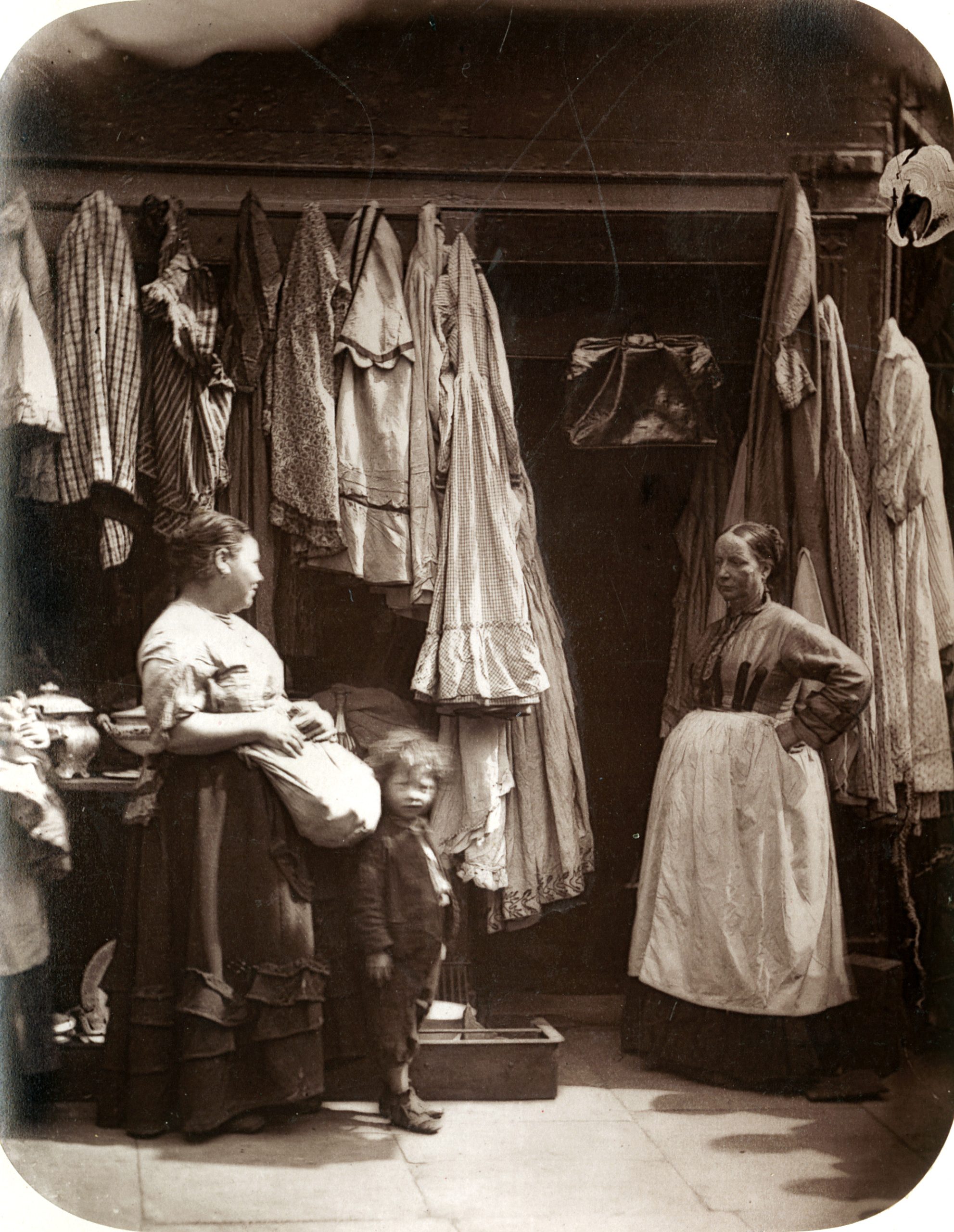 London’s poor and the second-hand trade: women and a child outside an old clothes shop in Lumber Court, 1877